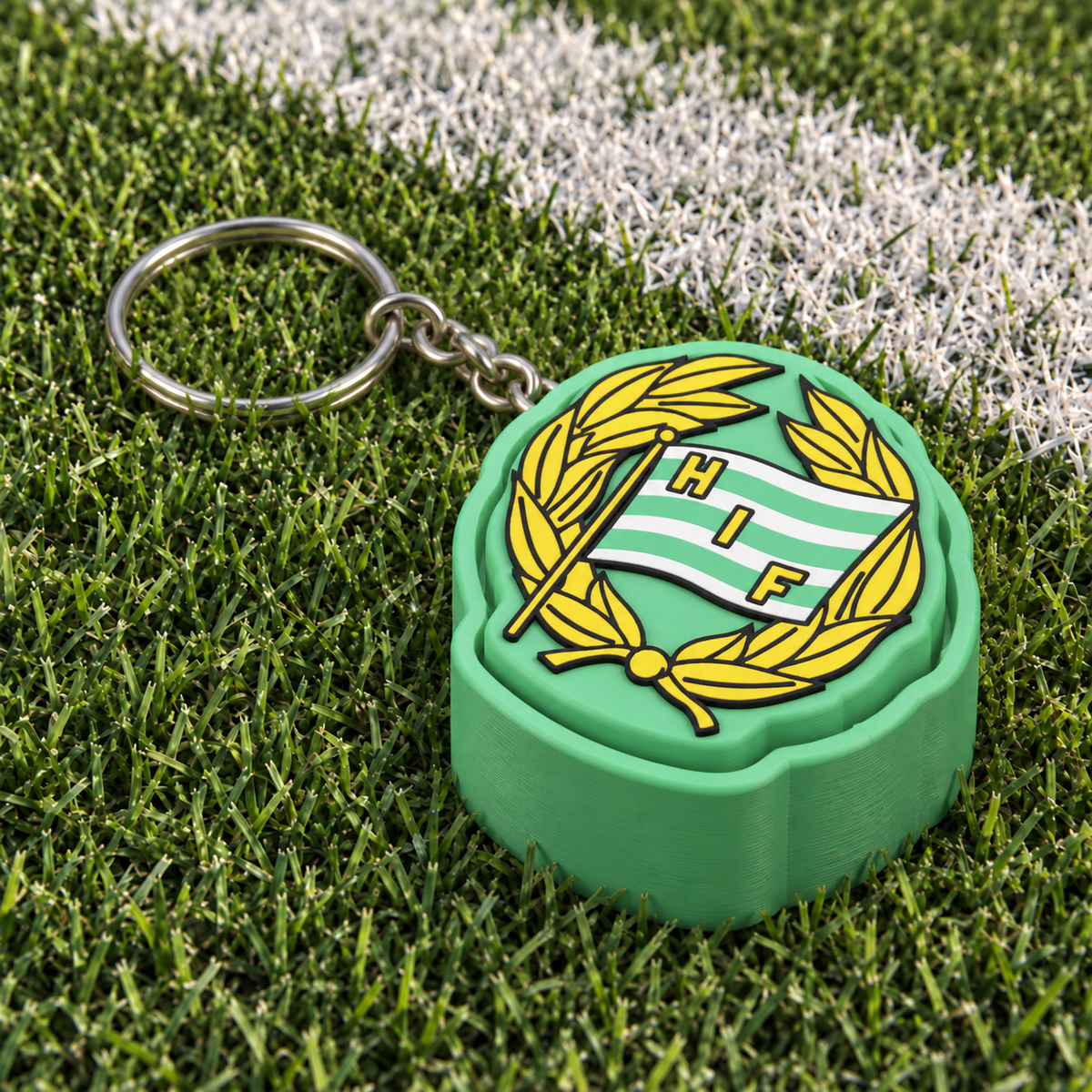 Hammarby clicker keychain with a green base, yellow laurel wreath and green-and-white HIF flag, resting on football turf with a short metal chain and silver split ring.