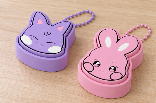 Purple SANdeoki and pink DDEONGbyeoli clickers with matching ball chains extending behind the tops of their bases on a light wooden surface.