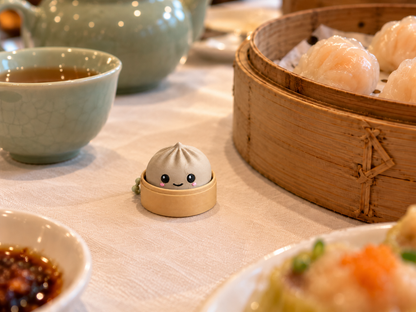 Bone white 3D-printed dumpling clicker keychain on a white linen dim sum table next to a bamboo steamer of har gow and a celadon teacup