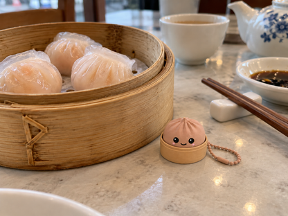 Latte brown 3D-printed dumpling clicker keychain on a dim sum table next to a bamboo steamer of har gow