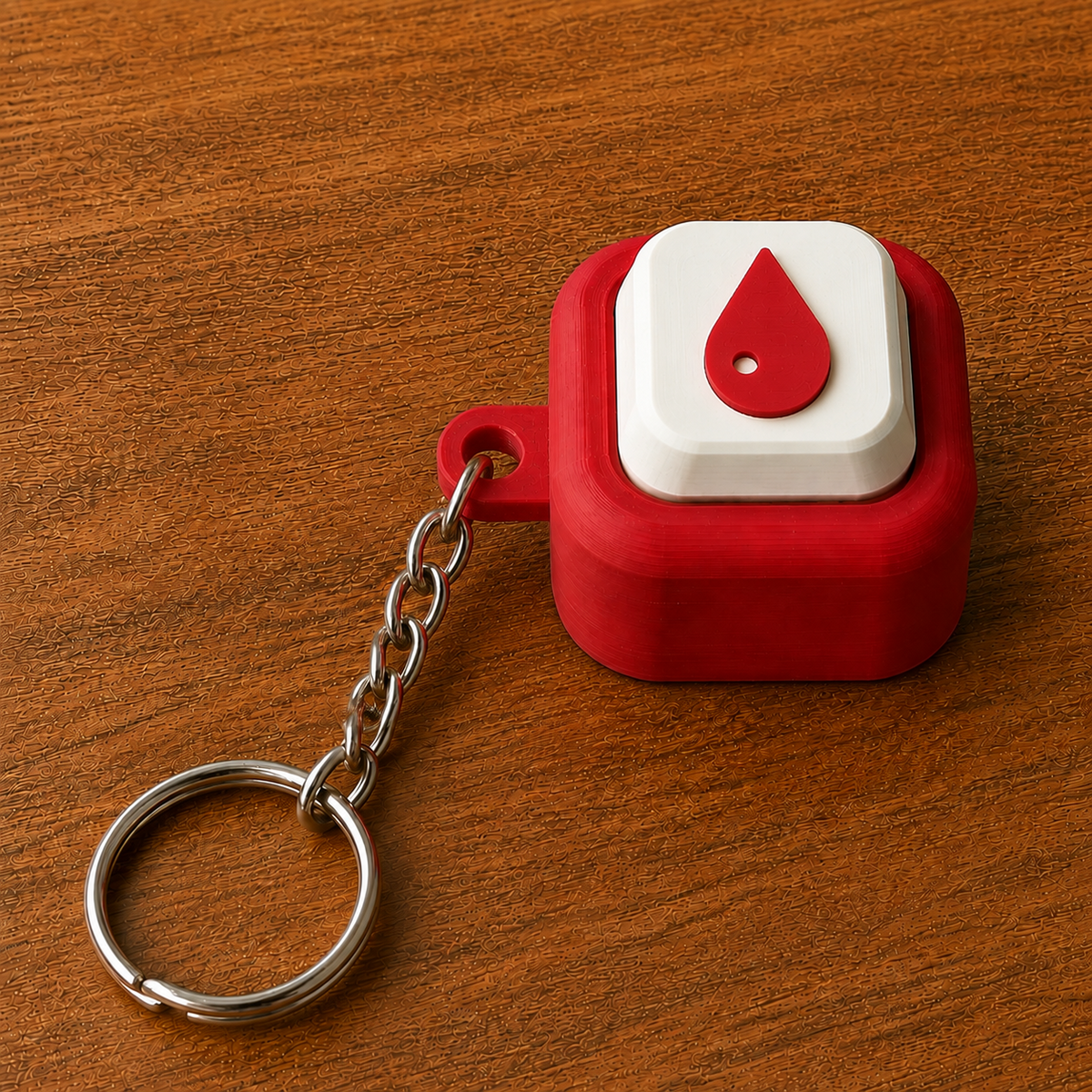 Red and white Hospital Clicker with a blood drop design, raised white button, side loop, metal chain and split ring on a wooden surface.