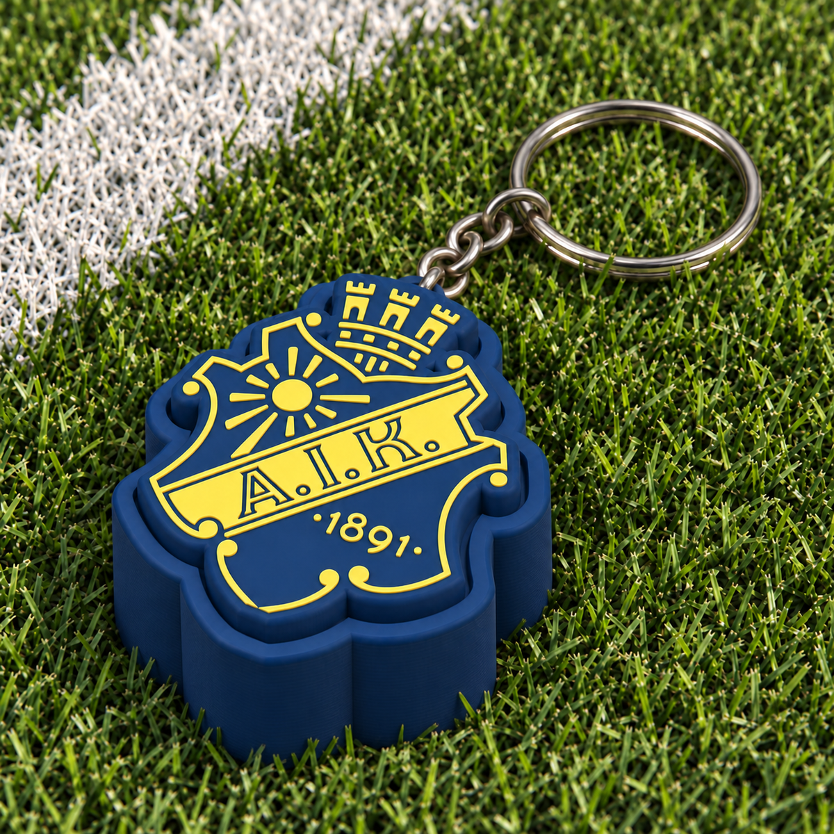 Navy and yellow AIK clicker keychain resting on football turf near a white pitch marking.