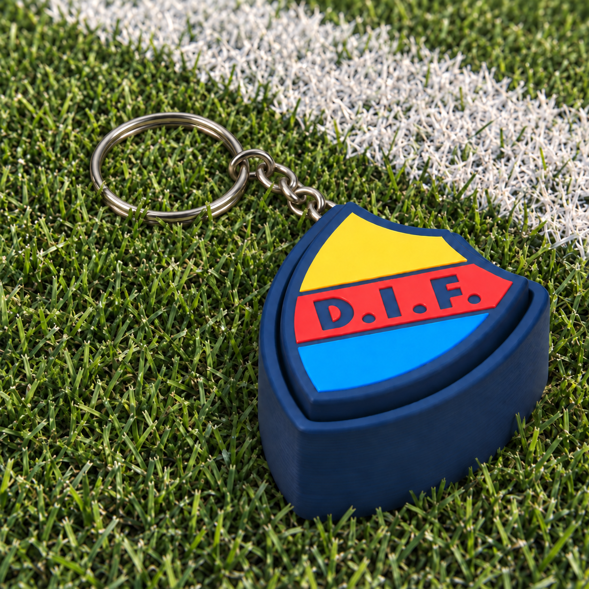 Djurgården clicker keychain in navy, yellow, red and blue on football turf beside a white line.
