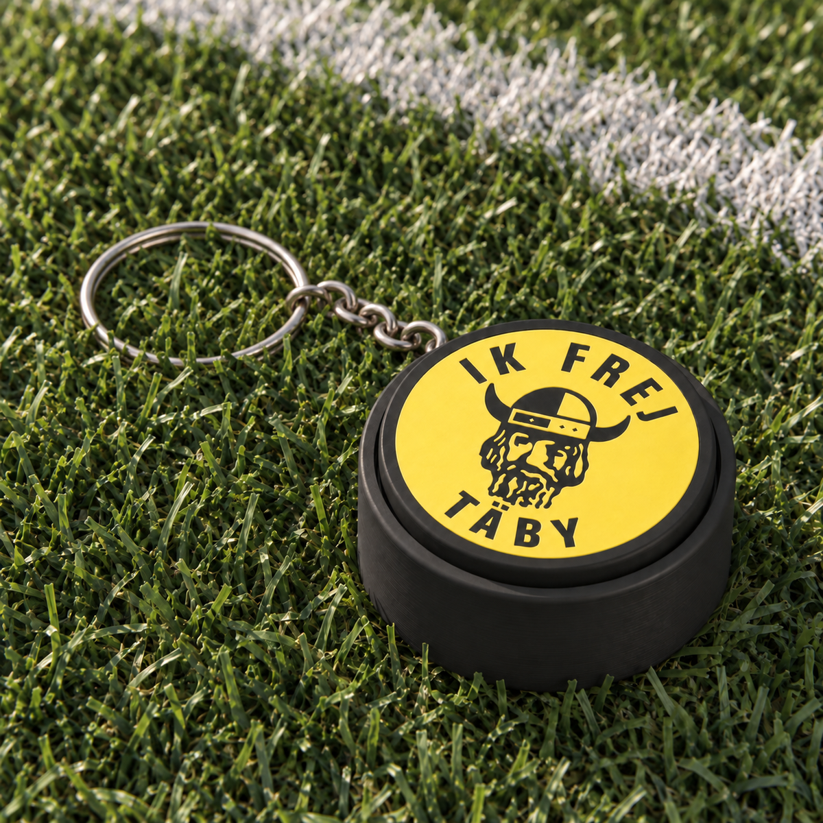 Round black and yellow IK Frej clicker with a Viking motif and silver keyring resting among football grass.