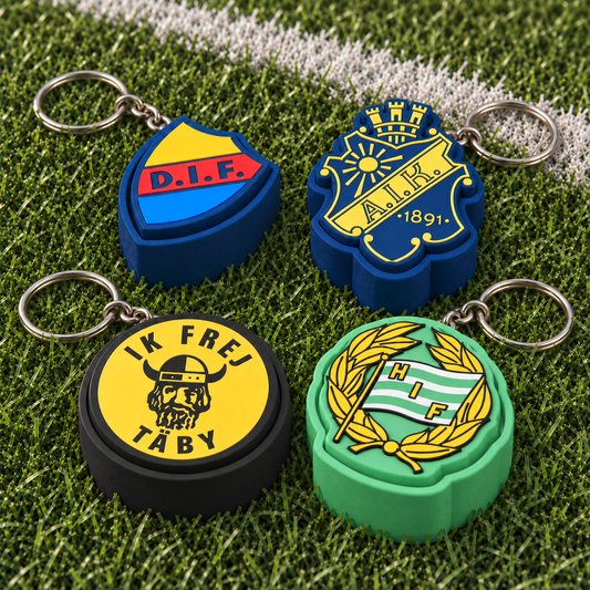 Four team clicker keychains on football turf: Djurgården and AIK behind, IK Frej and Hammarby in front, each with a short metal chain and silver keyring.